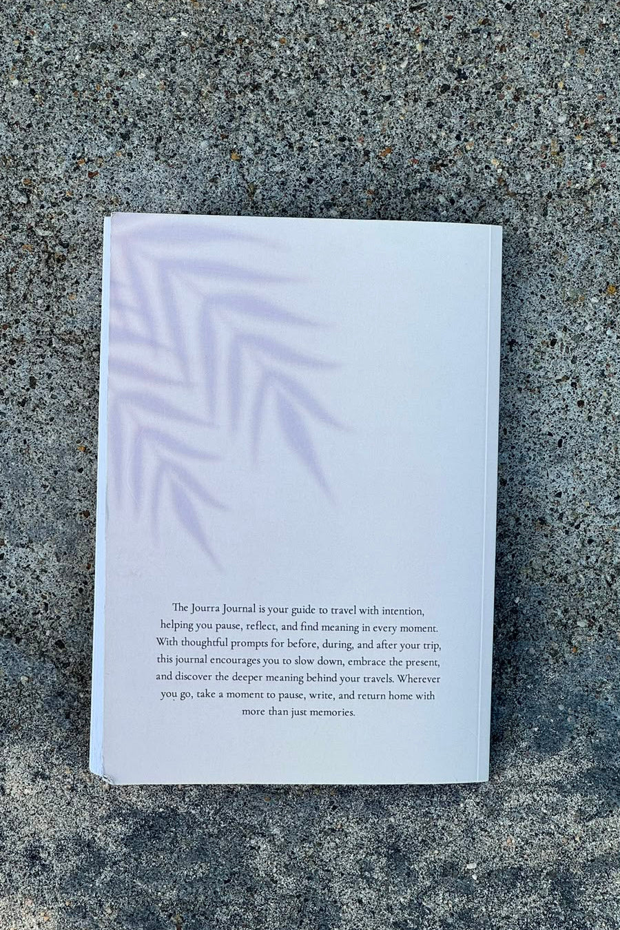 A white hardcover journal with a minimalist pale green leaf pattern on the cover and black printed text on the back, photographed from above on a textured gray concrete surface.