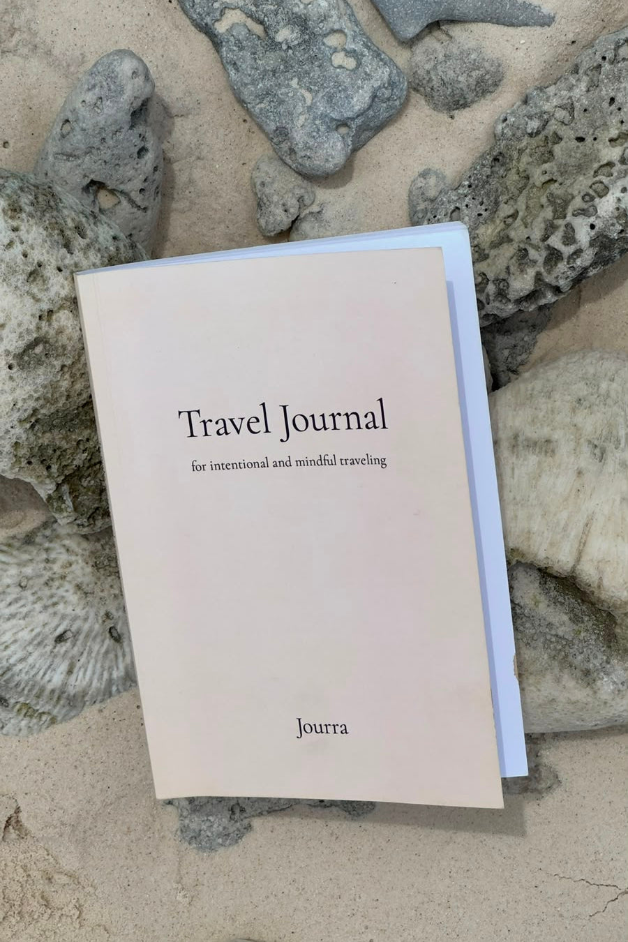 A cream-colored paper travel journal titled 'Travel Journal' with the brand name 'Jourra' printed on the cover, resting on a sandy surface surrounded by small gray rocks.