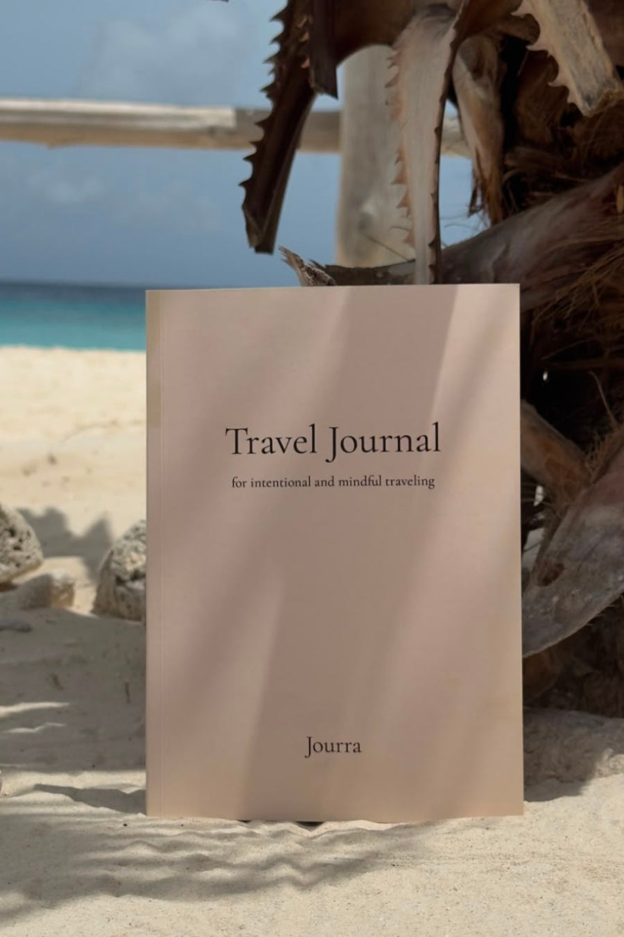 A beige travel journal standing upright on a sandy beach, with a tropical beach scene and a large dried palm frond in the background. The cover reads 'Travel Journal' and shows the brand name 'joura' near the bottom.
