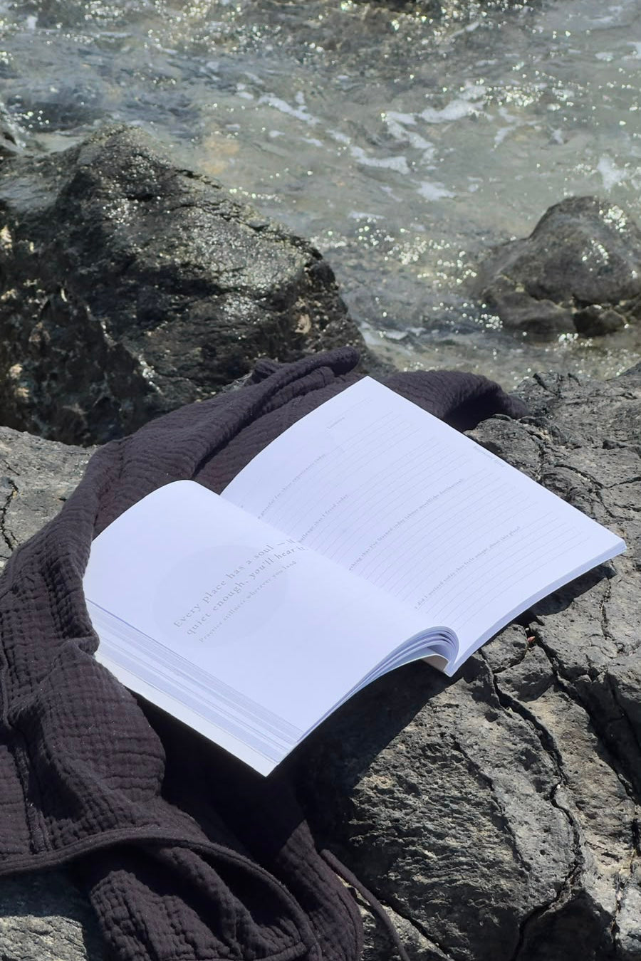 An open white journal lies on dark rocks by the water, with a dark knit blanket draped beside it and ocean waves visible in the background.