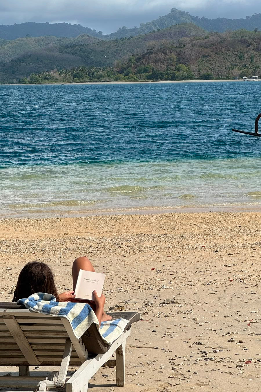 A person relaxes on a beach chair on a sandy beach, reading a book with the ocean and mountains in the background.